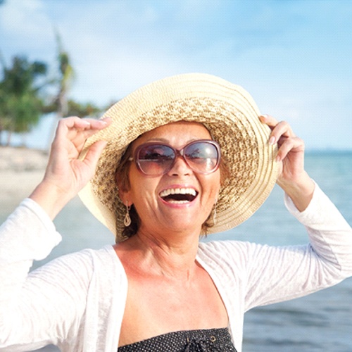 An older woman holding her hat while smiling on the beach after receiving implant dentures in Jacksonville