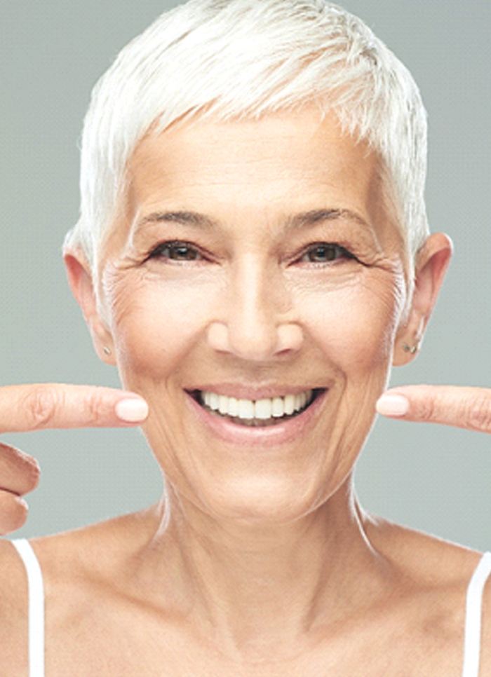 An older woman smiling and pointing to her new dentures in Jacksonville